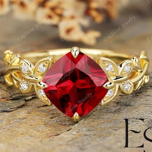 May include: A gold ring featuring a large, square-cut, vibrant red gemstone. The ring's band is adorned with delicate floral and leaf designs, accented with small, sparkling white stones. The ring is set against a natural, textured background.