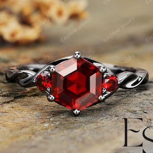 May include: A black gold engagement ring with a large red gemstone in a hexagonal setting. Two smaller red gemstones are set on either side of the main stone. The ring has a twisted band.