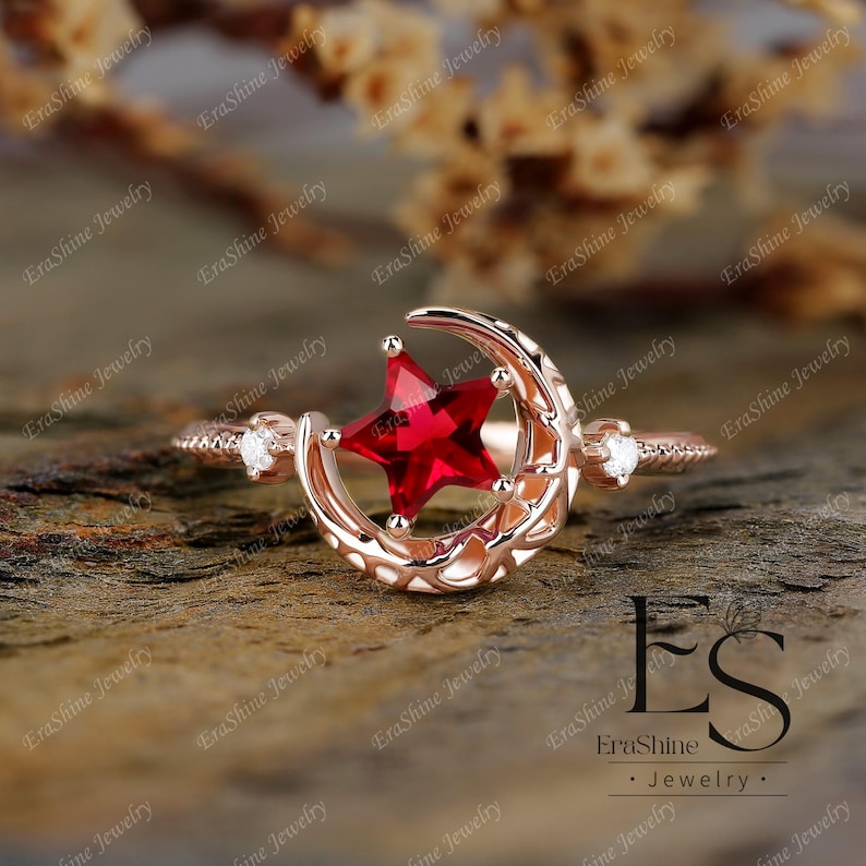 K&ouml;nnte beinhalten: Ein Ring aus Ros&eacute;gold mit einem Halbmond-Design und einem roten sternf&ouml;rmigen Edelstein. Der Ring hat zwei kleine klare Edelsteine auf der Band.