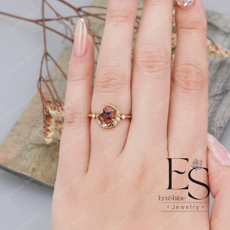 K&ouml;nnte beinhalten: Ein goldener Ring mit einem Halbmond-Design und einem roten Edelstein in der Mitte. Der Ring ist mit kleinen wei&szlig;en Edelsteinen auf beiden Seiten des roten Edelsteins besetzt. Der Ring befindet sich an einem Finger.