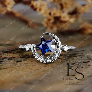 K&ouml;nnte beinhalten: Ein silberner Ring mit einem Halbmond-Design und einem blauen sternf&ouml;rmigen Edelstein. Der Ring hat zwei kleine klare Edelsteine auf der Band.
