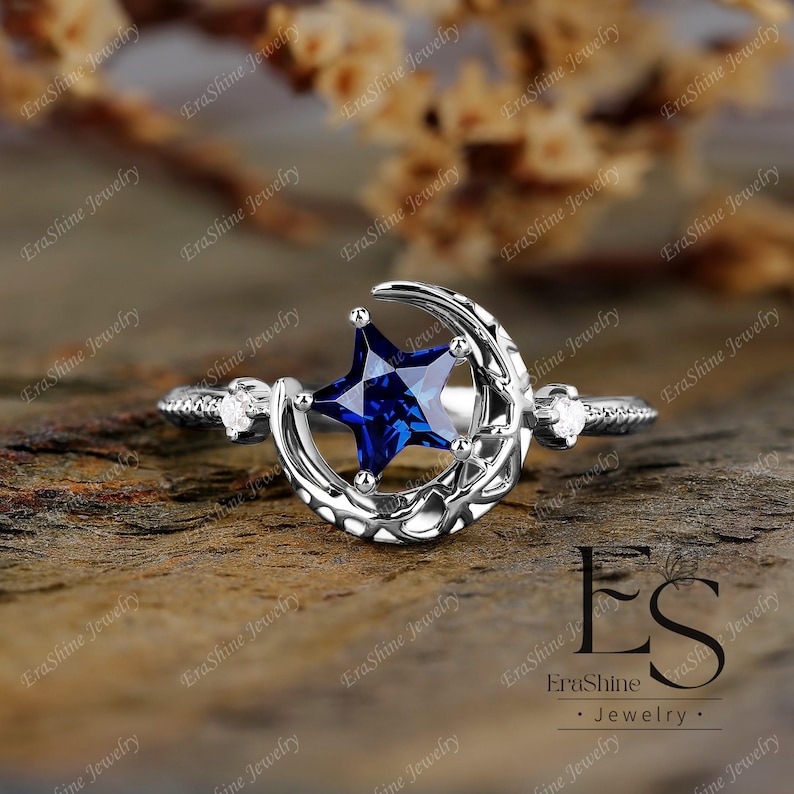 K&ouml;nnte beinhalten: Ein silberner Ring mit einem Halbmond-Design und einem blauen sternf&ouml;rmigen Edelstein. Der Ring hat zwei kleine klare Edelsteine auf der Band.