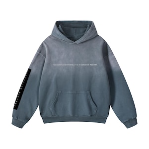 May include: A blue-grey hoodie with a front pocket and a drawstring hood. The text "TO ALLOW FLOW NATURALLY IS TO CONCEIVE MASTERY" is printed across the chest. A black rectangle on the left sleeve displays vertical text.