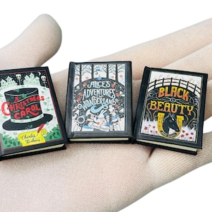 Peut inclure: Trois livres miniatures avec des couvertures noires sont présentés. Les livres sont intitulés "Christmas Carol", "Alice au pays des merveilles" et "Black Beauty". Les livres sont tenus dans une main gantée.