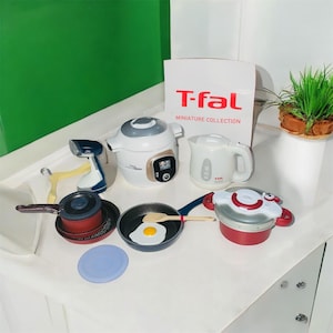 Op de afbeelding: Een miniatuurkeukenset met een witte en grijze elektrische snelkookpan, een rode en witte snelkookpan, een rode pan, een zwarte koekenpan met een gebakken ei, een witte elektrische waterkoker en een blauwe en witte handstoompan. De set is gemarkeerd met "T-fal Miniature Collection".