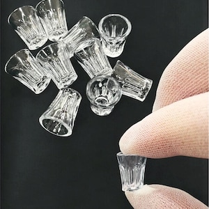 May include: A selection of small, clear shot glasses with a traditional design. The glasses are made of transparent glass and are scattered on a dark surface. One glass is held by a gloved hand.