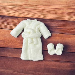 May include: A miniature white terry cloth bathrobe with a matching belt and a pair of slippers. The items are arranged on a wooden surface with a warm, brown tone. The bathrobe has long sleeves.