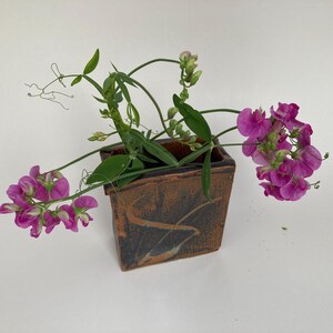 May include: A square, brown ceramic vase with a dark, abstract design holds vibrant pink flowers and green foliage. The flowers are in full bloom, with delicate petals and a rich color. The vase is placed on a white surface.