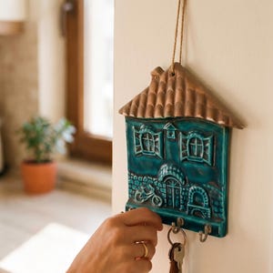 May include: A wall-mounted ceramic key holder shaped like a house with a brown roof and a teal facade. The house features embossed details including two windows, a front door, a bicycle, and three metal hooks at the bottom for hanging keys.