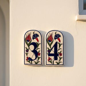 May include: Two ceramic house number plaques, displaying the numbers 3 and 4. Each plaque features a white background with a blue border and a floral design in red, blue, and green. The numbers are in a dark blue colour.
