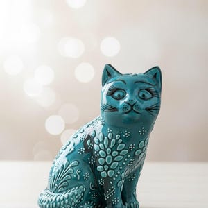 Może przedstawiać: Turkusowa ceramiczna figurka kota ze skomplikowanymi wzorami kwiatowymi i kropkowymi. Kot ma duże oczy i uśmiechniętą minę. Figurka siedzi na jasnej powierzchni, z rozmytym tłem z miękkich białych kół.