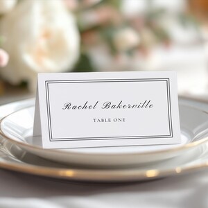 Può includere: Un segnaposto bianco con un bordo nero e il testo "Rachel Bakerville" e "Tavolo Uno" stampato in nero.