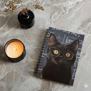 May include: A spiral-bound notebook featuring a black cat wearing glasses, set against a chalkboard background. A lit candle in a black container and a small black vase with dried stems are also present on a marble surface.