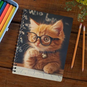 May include: Spiral-bound notebook featuring a ginger kitten wearing glasses and a bell collar. The kitten is depicted on a dark background with faint text. The notebook is open, with a partially visible page. Pencils and colored markers are also in the image.