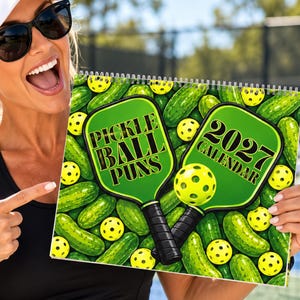 May include: A 2027 calendar featuring a graphic design of two green pickleball paddles and yellow perforated balls set against a background of green pickles. The text on the paddles reads Pickle Ball Puns and 2027 Calendar in black font.