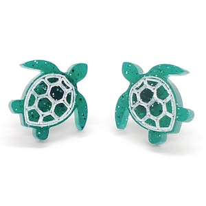 May include: A pair of green glitter turtle stud earrings with a silver outline. The turtles have a detailed shell design.