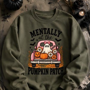 Könnte beinhalten: Ein olivgrüner Rundhals-Pullover mit einer Grafik eines rosa Pickups, der ein Gespenst mit rosa Schleife, orangefarbene Kürbisse und weiße Blumen geladen hat. Der Text lautet MENTALLY at the PUMPKIN PATCH, umgeben von kleinen schwarzen Fledermäusen.