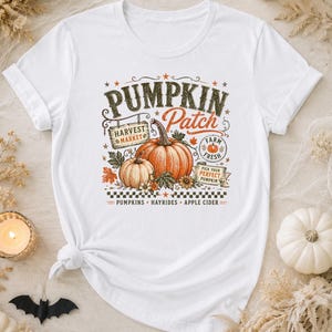 May include: A white short-sleeve t-shirt featuring a graphic print of two orange pumpkins, autumn leaves, and text that reads PUMPKIN Patch, HARVEST MARKET, FARM FRESH, PICK YOUR PERFECT PUMPKIN, PUMPKINS, HAYRIDES, and APPLE CIDER with a checkered border.