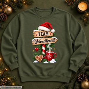 May include: An olive green sweatshirt featuring a graphic of a wooden signpost with a Santa hat, pine branches, a heart-shaped cookie, and a red mug with a snowflake. The sign reads "TEAM" and "Weihnachtsmarkt" in black text.