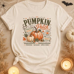 May include: A cream-colored t-shirt featuring a graphic print of two orange pumpkins with the text PUMPKIN PATCH, HARVEST MARKET, FARM FRESH, PICK YOUR PERFECT PUMPKIN, PUMPKINS, HAYRIDES, and APPLE CIDER, accented with autumn leaves and flowers.