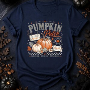 May include: A navy blue t-shirt featuring a graphic print with the text PUMPKIN PATCH, HARVEST MARKET, FARM FRESH, and PICK YOUR PERFECT PUMPKIN. The design includes illustrations of two pumpkins, autumn leaves, flowers, and the words PUMPKINS, HAYRIDES, APPLE CIDER.