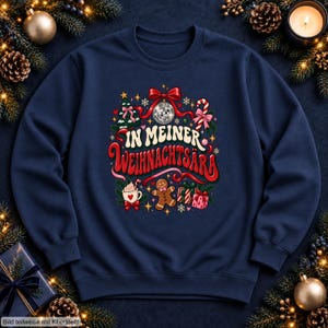 May include: A navy blue sweatshirt featuring a graphic print with the text "IN MEINER WEIHNACHTSÄRA" surrounded by a disco ball, a Christmas tree, a candy cane, a mug of hot cocoa, a gingerbread man, and wrapped gift boxes.