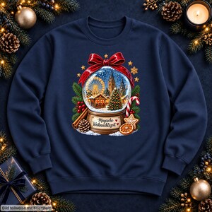 May include: A navy blue sweatshirt featuring a graphic of a snow globe topped with a red bow. Inside the globe is a Christmas market scene with a Ferris wheel and tree. The base reads Magische Weihnachtszeit, surrounded by pine, candy cane, and orange.