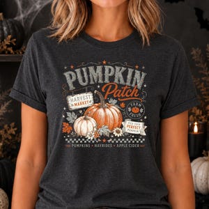 May include: A dark charcoal gray t-shirt featuring a graphic print with the text PUMPKIN PATCH, HARVEST MARKET, FARM FRESH, and PICK YOUR PERFECT PUMPKIN. The design includes two pumpkins, autumn leaves, flowers, and the words PUMPKINS, HAYRIDES, APPLE CIDER.