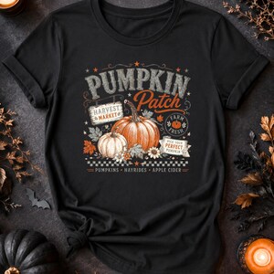 May include: A black t-shirt featuring a graphic print with the text PUMPKIN PATCH, HARVEST MARKET, FARM FRESH, and PICK YOUR PERFECT PUMPKIN. The design includes illustrations of two pumpkins, autumn leaves, flowers, and a checkered border pattern.
