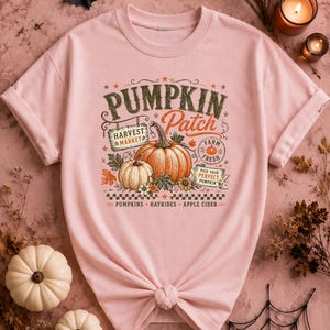 May include: A light pink t-shirt featuring a graphic print with the text PUMPKIN PATCH, HARVEST MARKET, FARM FRESH, PICK YOUR PERFECT PUMPKIN, and PUMPKINS HAYRIDES APPLE CIDER. The design includes illustrations of orange and white pumpkins.