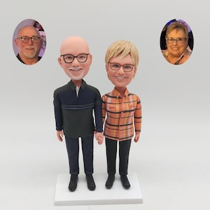 Könnte beinhalten: Eine personalisierte Wackelkopf-Figur eines Paares auf einem weißen Sockel. Die linke Figur ist eine glatzköpfige Person mit Brille, die ein dunkelblaues Hemd und eine Hose trägt. Die rechte Figur hat kurzes blondes Haar, eine Brille und ein orangefarbenes Karohemd.