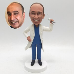 May include: A figurine of a smiling doctor in a white lab coat, blue scrubs, and black shoes, holding dental tools. A photo of a man's face is in the upper left corner. The figurine stands on a white circular base.
