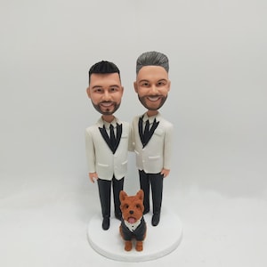 Könnte beinhalten: Zwei Bräutigame in weißen Smokingjacken mit schwarzen Fliegen und schwarzen Hosen stehen nebeneinander. Ein kleiner brauner Hund mit einer schwarzen Fliege steht vor ihnen. Die Bräutigame und der Hund sind Figuren.