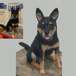 May include: A detailed sculpture of a dog, predominantly black with tan markings, sitting on a round, textured surface. The dog has alert ears and a slightly open mouth. An inset image shows the dog in a similar pose.