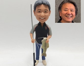 May include: A figurine of a smiling person holding a fishing rod and a fish. The figure wears a black and gray polo shirt, blue pants, and beige shoes. A photo of a smiling man is in the background. The figurine is on a white base.