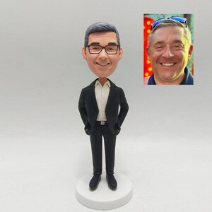 May include: A custom bobblehead figurine of a man wearing a black suit, white shirt, and glasses. The figurine is standing on a white circular base. A small photo of the man is in the upper right corner.