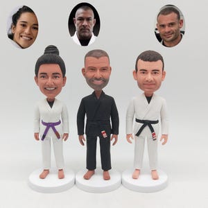 Puede incluir: Tres figuras cabezonas personalizadas que representan a personas con uniformes de artes marciales. La figura de la izquierda lleva un gi blanco con cinturón morado, la del centro un gi negro con cinturón negro y la de la derecha un gi blanco.