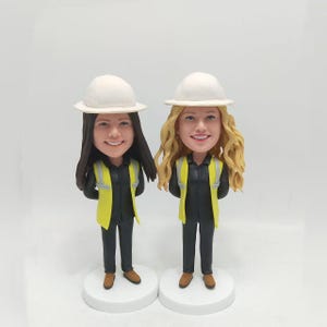 May include: Two custom-made figurines of people wearing white hard hats and yellow safety vests. The figurines are standing on white bases.