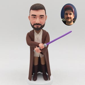 May include: A custom bobblehead figurine featuring a bearded man with short brown hair, wearing a long brown robe over a cream-coloured tunic and dark brown boots. The figure holds a purple lightsaber in both hands, with a small circular photo inset.