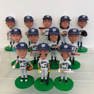 Könnte beinhalten: Eine Sammlung von Baseball-Wackelkopf-Figuren, jede mit einem Spieler in einer weiß-blauen Nadelstreifenuniform und einer blauen Kappe mit einem "W"-Logo. Die Figuren stehen auf grünen, kreisförmigen Sockeln. Einige halten Schläger, Handschuhe oder Basebälle.