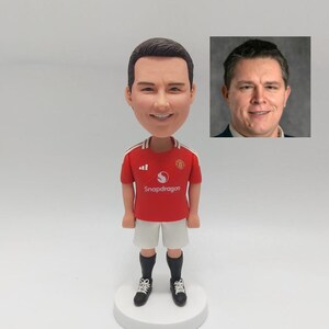 May include: A custom bobblehead figurine of a person wearing a red and white soccer jersey with the text "Snapdragon" on the front. The jersey has the Manchester United logo on the right side of the chest. The figurine is standing on a white base.