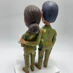 May include: A pair of custom-made figurines of a couple in military uniform. The figurines are standing close together, with the woman's arm around the man's waist. They are both wearing camouflage patterned uniforms.