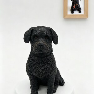 Könnte beinhalten: Eine schwarze Hundefigur mit strukturiertem Fell, die auf einem weißen runden Sockel sitzt. Über der Figur befindet sich ein heller Holzrahmen mit einem Foto desselben schwarzen Hundes, der ein Halsband mit roter Marke trägt, vor weißem Hintergrund.