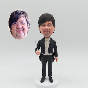 Könnte beinhalten: Eine personalisierte Wackelkopf-Figur eines Mannes im Smoking, der einen Taktstock hält. Die Figur hat dunkles Haar, eine Brille und eine Fliege. Ein ovales Foto eines Mannes mit Brille befindet sich neben der Figur. Die Figur steht auf einem weißen Sockel.