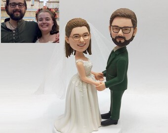 May include: A custom wedding cake topper featuring a bride in a white gown and a groom in a green suit, holding hands. The bride wears a veil and glasses. The groom has a beard and glasses. A small photo of the couple is in the background.