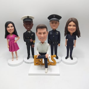 Op de afbeelding: Vijf gepersonaliseerde bobbleheads, elk met een uniek beroep. Een vrouw in een roze jurk, een man in een marineuniform, een man in een wit overhemd en blauwe jeans die op een gouden 'BOSS'-bord zit, een man in een politieuniform en een vrouw in een blauw uniform.