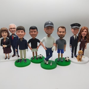 Op de afbeelding: Een verzameling gepersonaliseerde bobblehead-beeldjes, elk met een persoon in verschillende outfits en poses. De beeldjes staan op groene of witte bases, sommige met rekwisieten zoals golfclubs, voetballen en een hond.