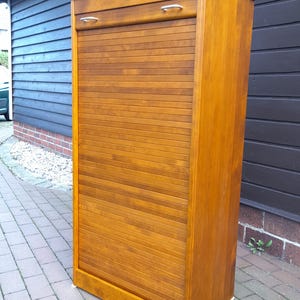 Könnte beinhalten: Ein hoher Holzschrank mit einer Rollladen-Tür. Der Schrank ist hellbraun und hat einen kleinen Metallgriff an der oberen Schublade.