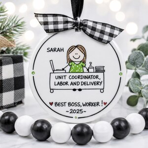 May include: A round white ceramic ornament featuring a black and white buffalo plaid ribbon. The design shows a cartoon figure in a green shirt behind a desk labeled UNIT COORDINATOR, LABOR AND DELIVERY, with the text SARAH, BEST BOSS, WORKER!, and 2025.