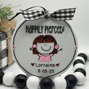 May include: A white ceramic ornament with the text "Happily Pierced!" and a cartoon girl illustration. The ornament has a black and white checkered bow and the name "Lorraine" with a date. A black and white bead garland is in the foreground.